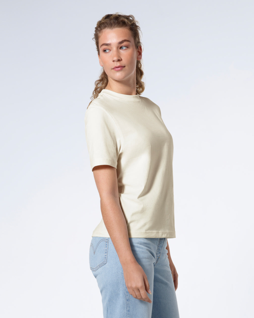 A woman facing side on looking back, modelling a plain ecru short sleeve organic cotton t shirt.