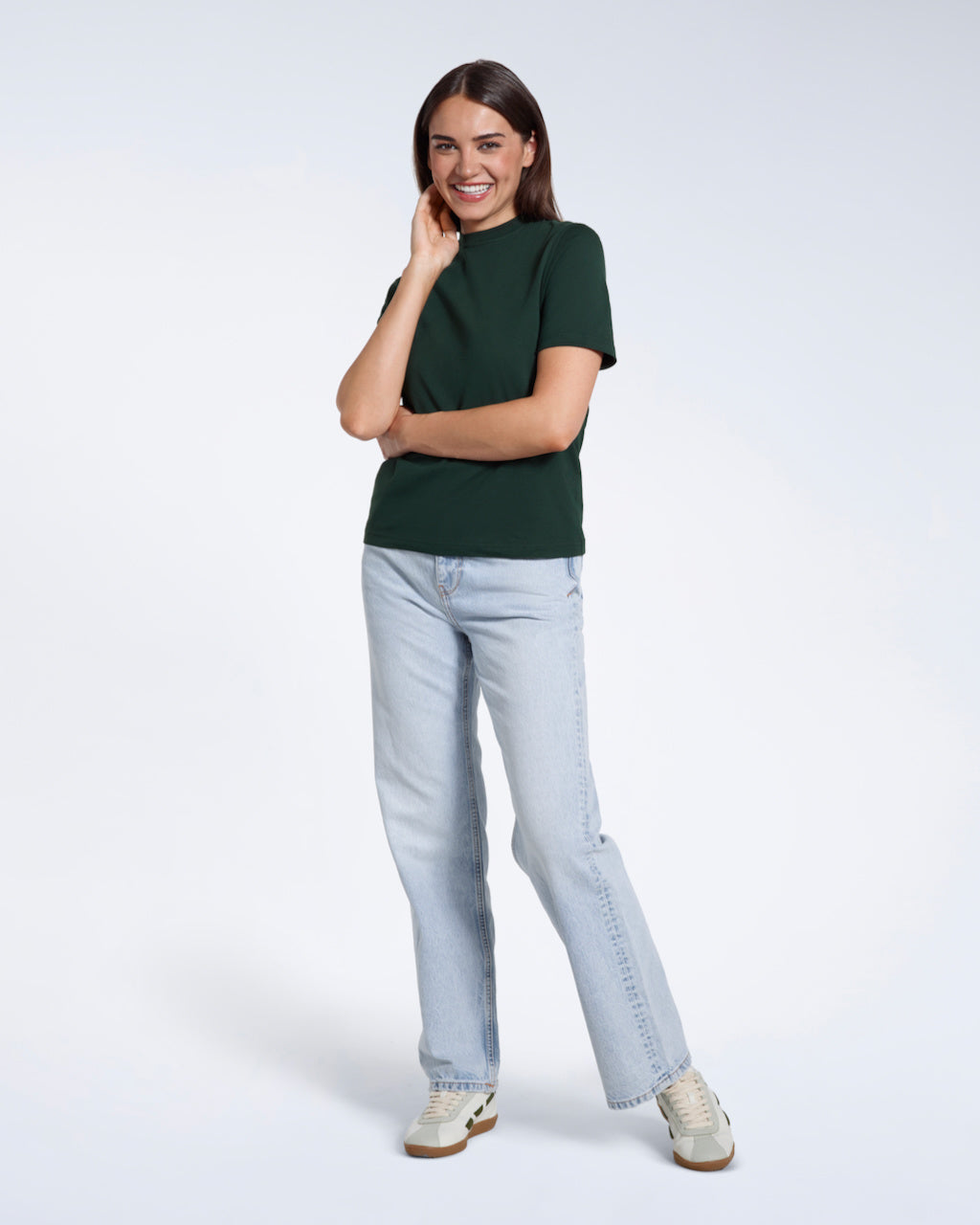 A full body photo of a woman touching her face smiling, modelling a plain green short sleeve organic cotton t shirt.