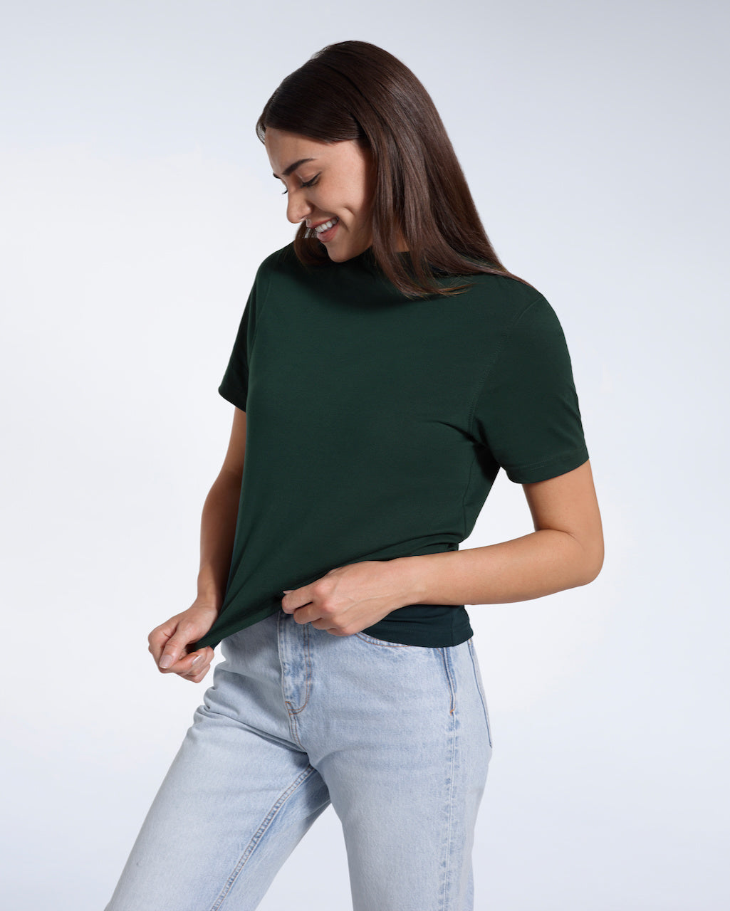 A woman looking down smiling and touching the bottom of her plain green short sleeve organic cotton t shirt.