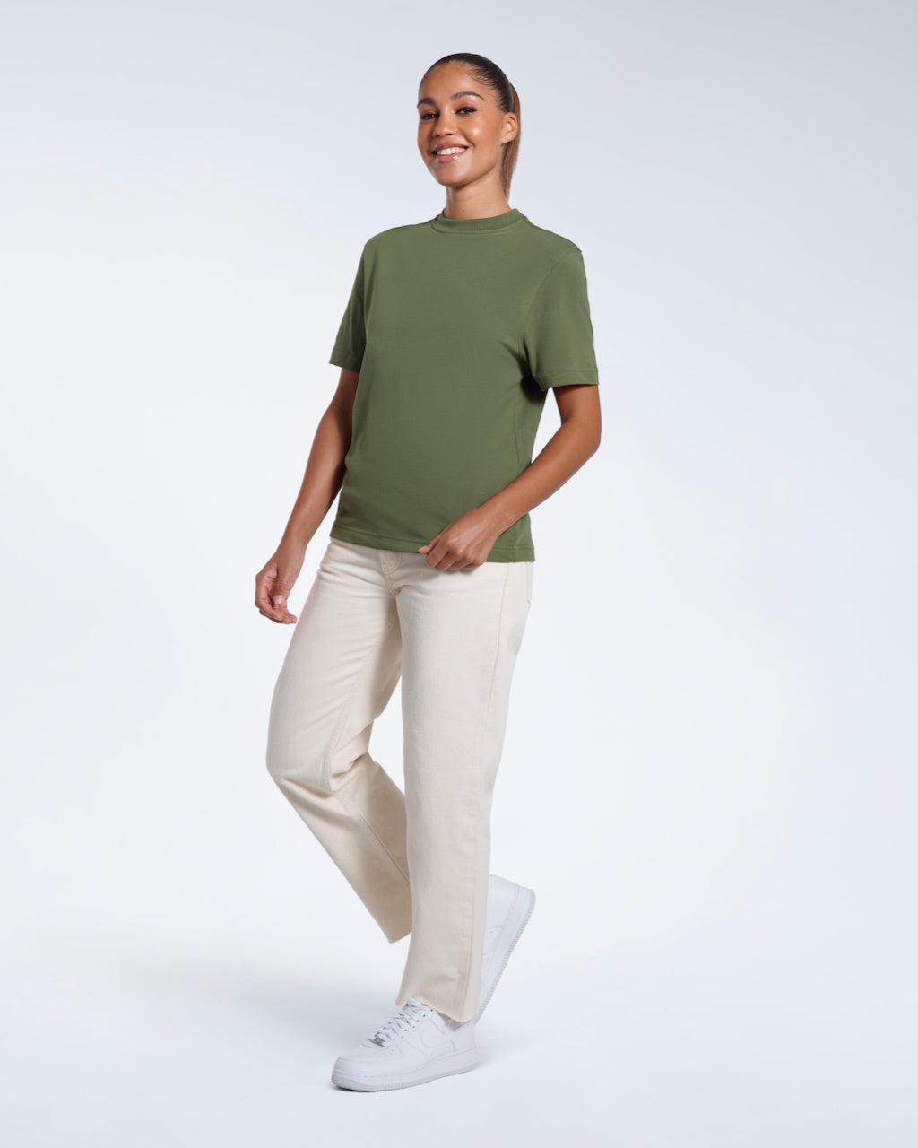A full body photo of a woman walking and smiling, modelling a plain khaki green short sleeve organic cotton t shirt.