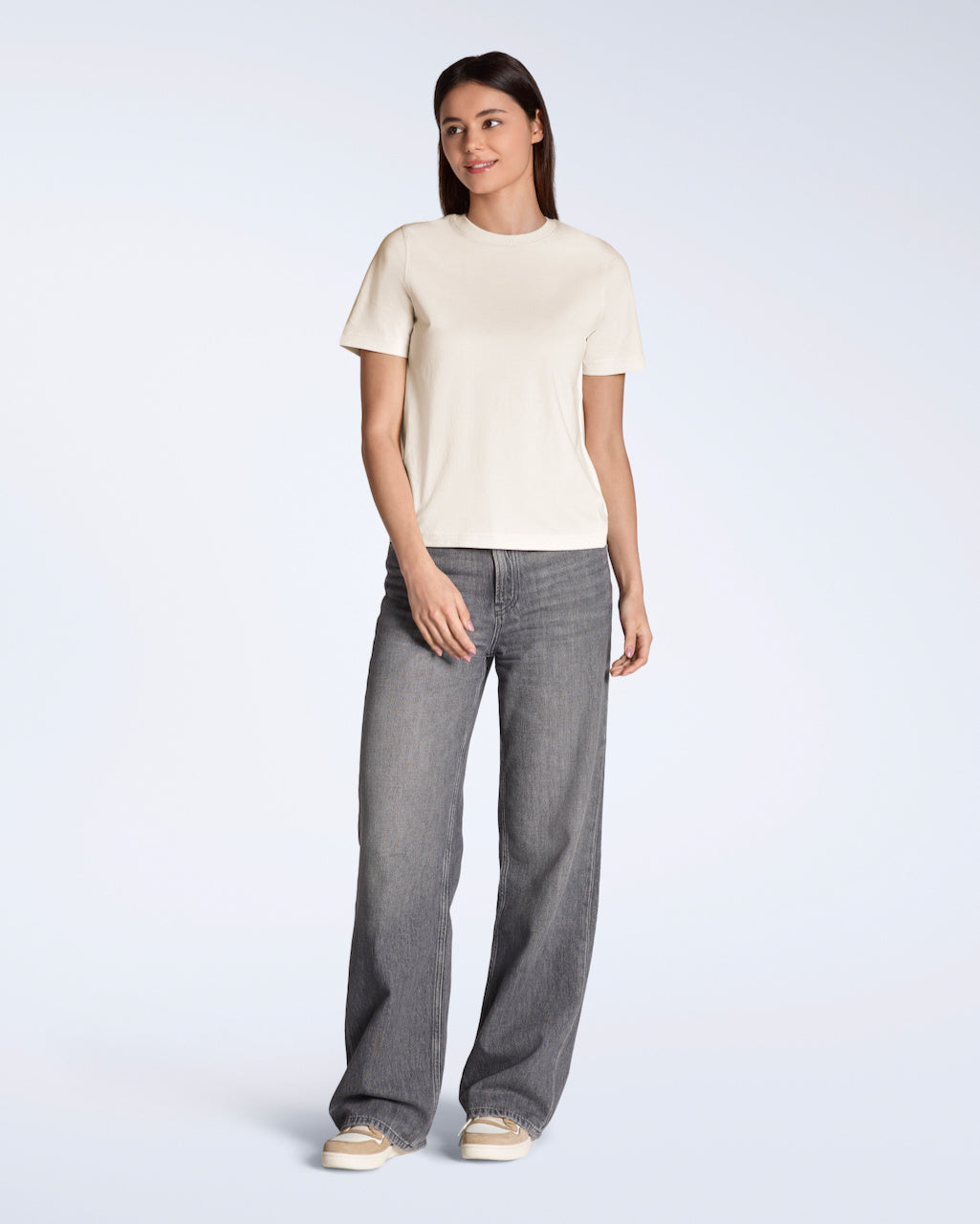 A full body photo of a woman smiling, modelling a plain natural coloured, organic cotton t shirt with a pair of grey jeans.