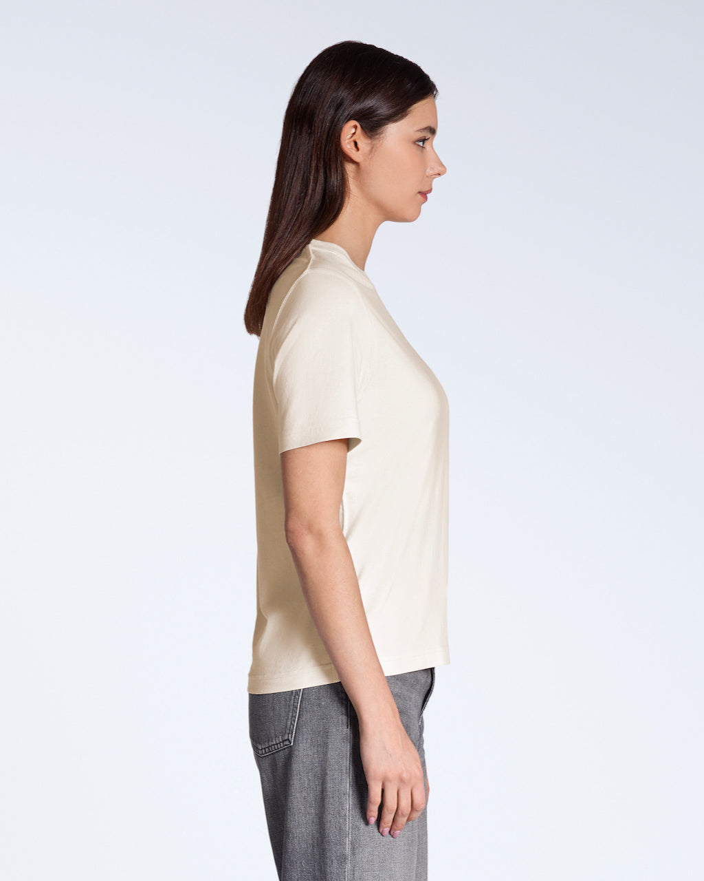 A woman facing side on, modelling a plain natural coloured, organic cotton t shirt with a pair of grey jeans.