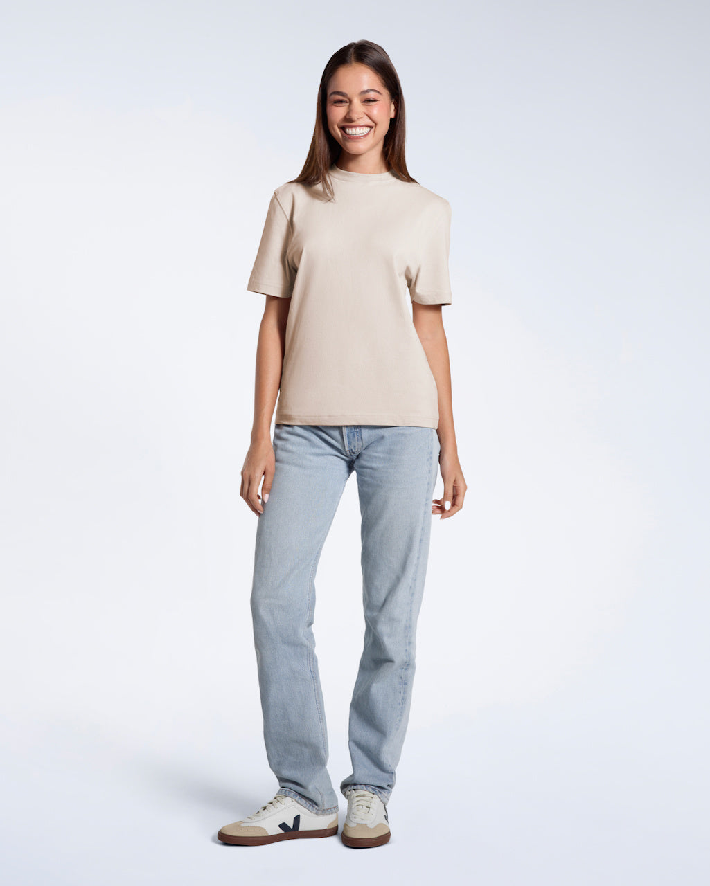 A full body photo of a woman smiling with her arms at her sides, modelling a plain stone coloured short sleeve organic cotton t shirt.