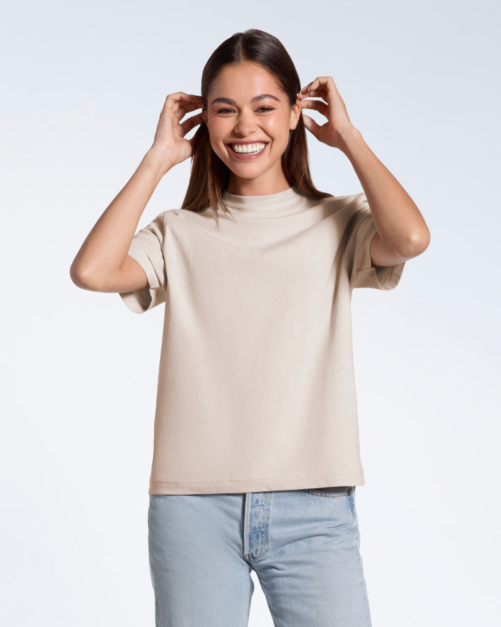 A woman smiling tucking her hair behind her ears, modelling a plain stone coloured short sleeve organic cotton t shirt.