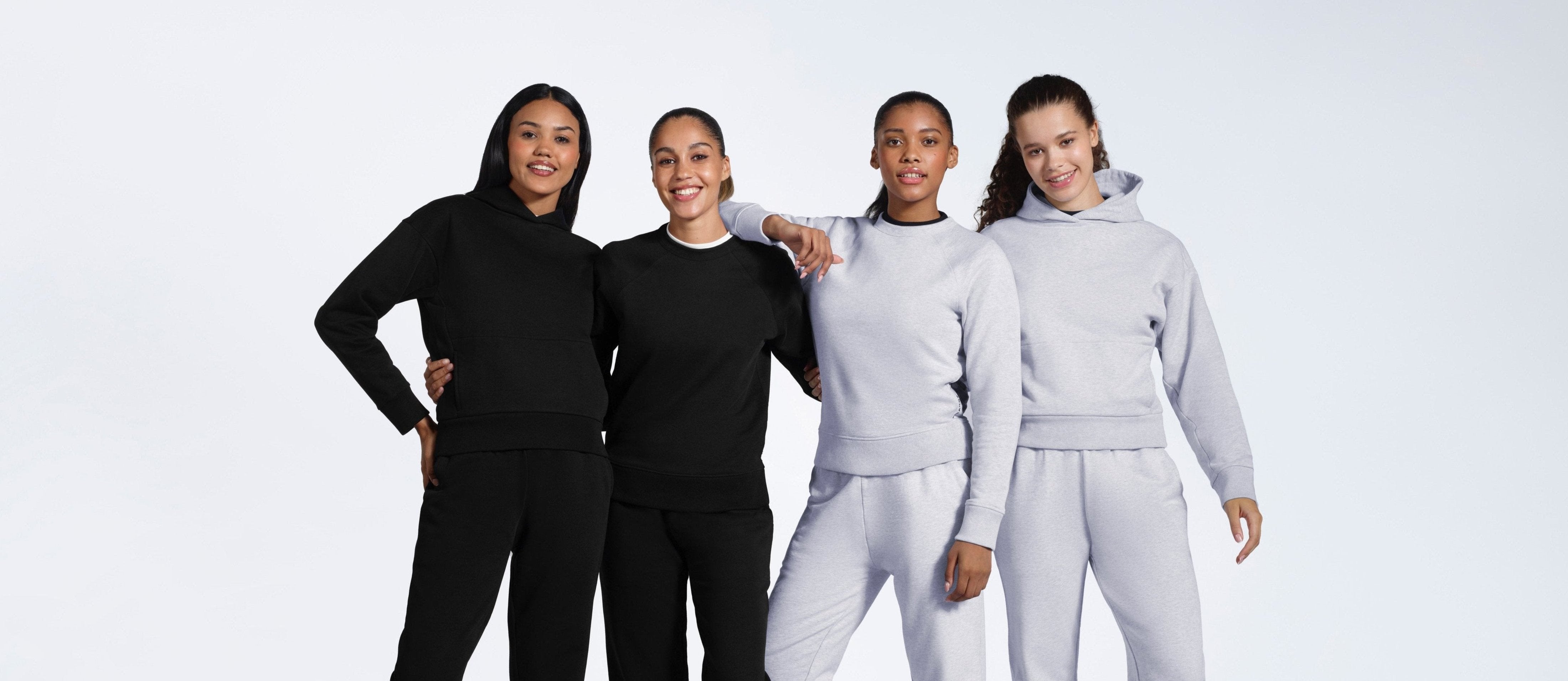 Four women smiling, modelling plain black and grey matching heavyweight organic cotton tracksuits.