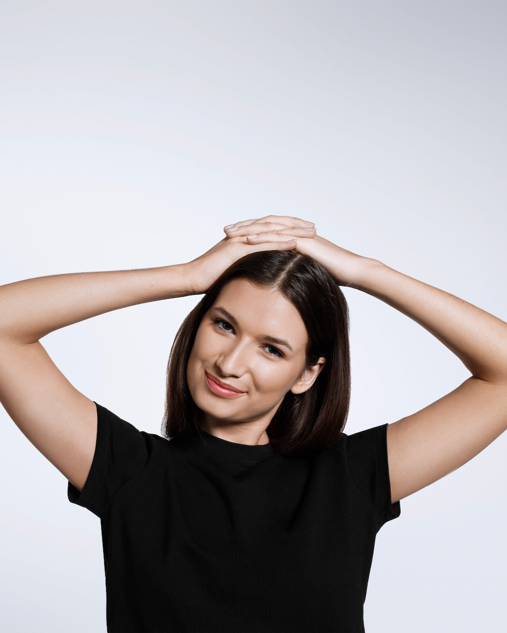 A woman smiling into the camera with her hands on her head, modelling a plain black short sleeve organic cotton t shirt.