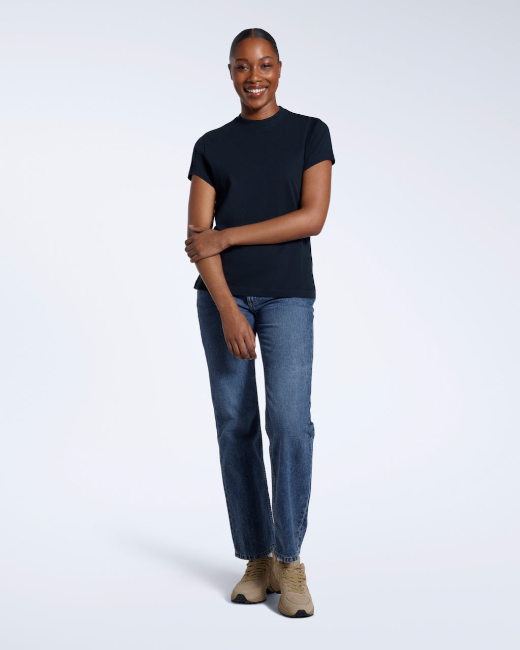 A full body photo of a woman smiling touching her arm, modelling a plain navy short sleeve organic cotton t shirt.