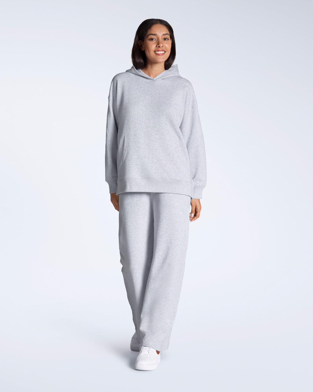 A woman smiling into the camera, modelling a matching grey oversized, organic cotton hoodie with a pair of wide leg joggers.