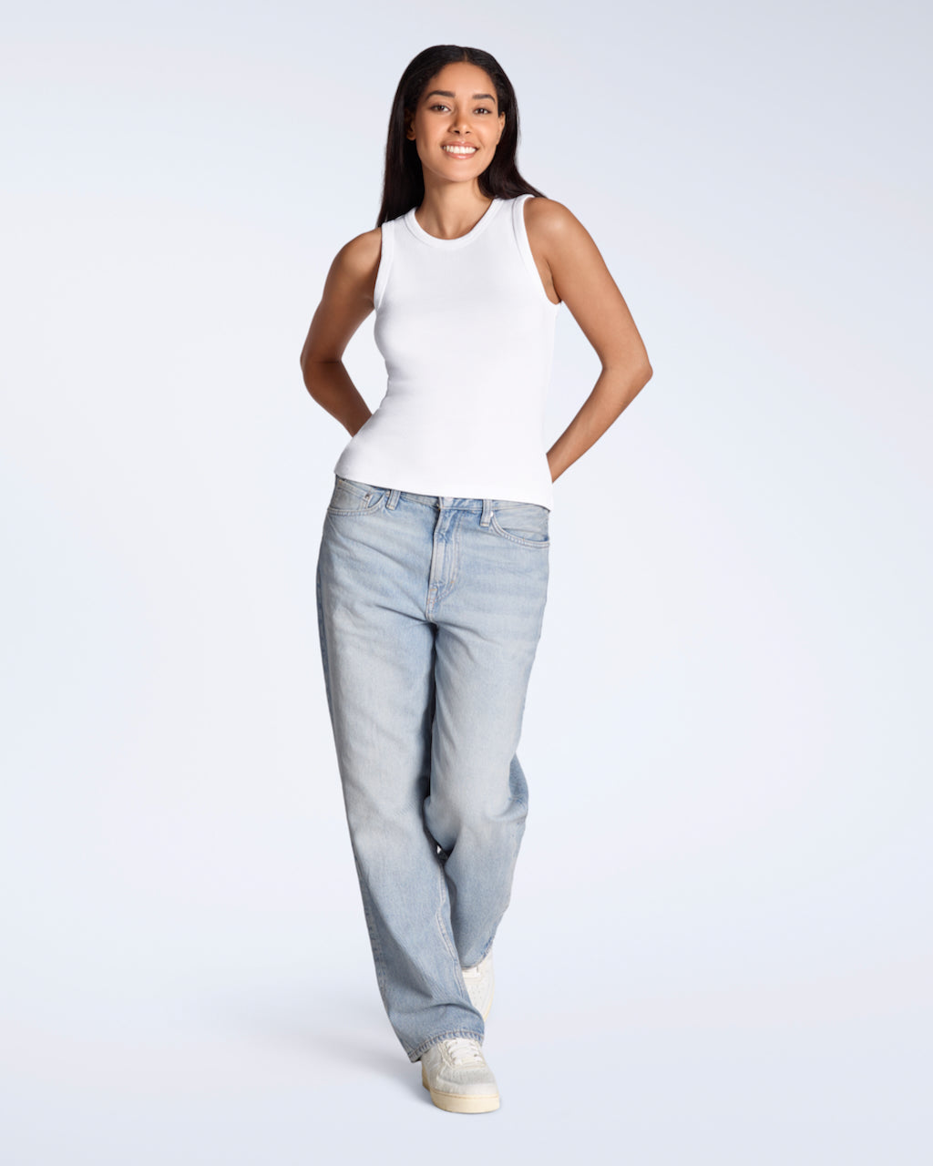 A full body photo of a woman smiling into the camera, modelling a plain white, organic cotton tank top with a pair of jeans.