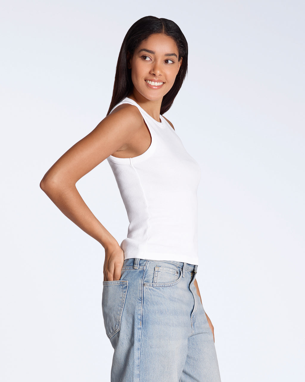 A woman smiling, facing side on with her hand in her back pocket, modelling a plain white, organic cotton tank top.