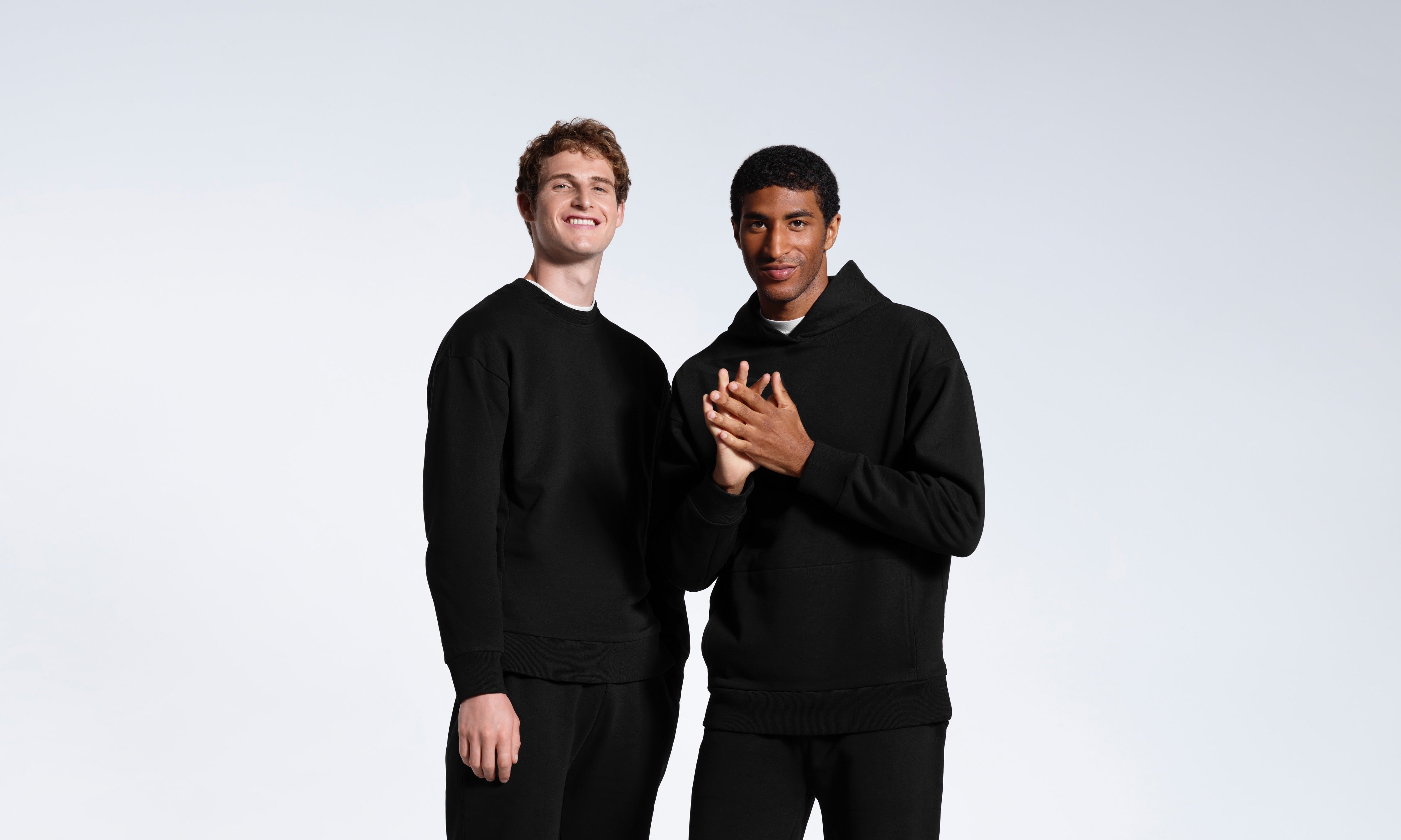 Two men in a plain black tracksuit set, smiling against a grey background.