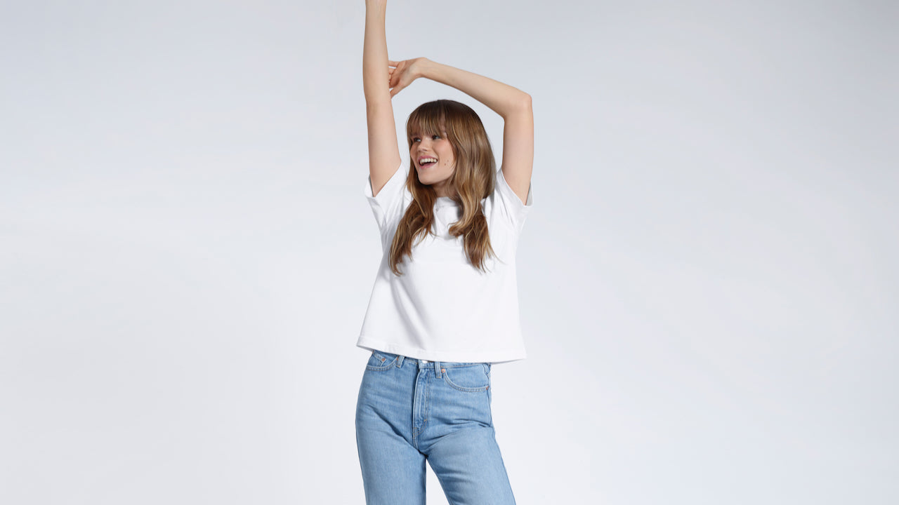 A woman with her arms above her head smiling, modelling a white boxy heavyweight organic cotton t shirt.