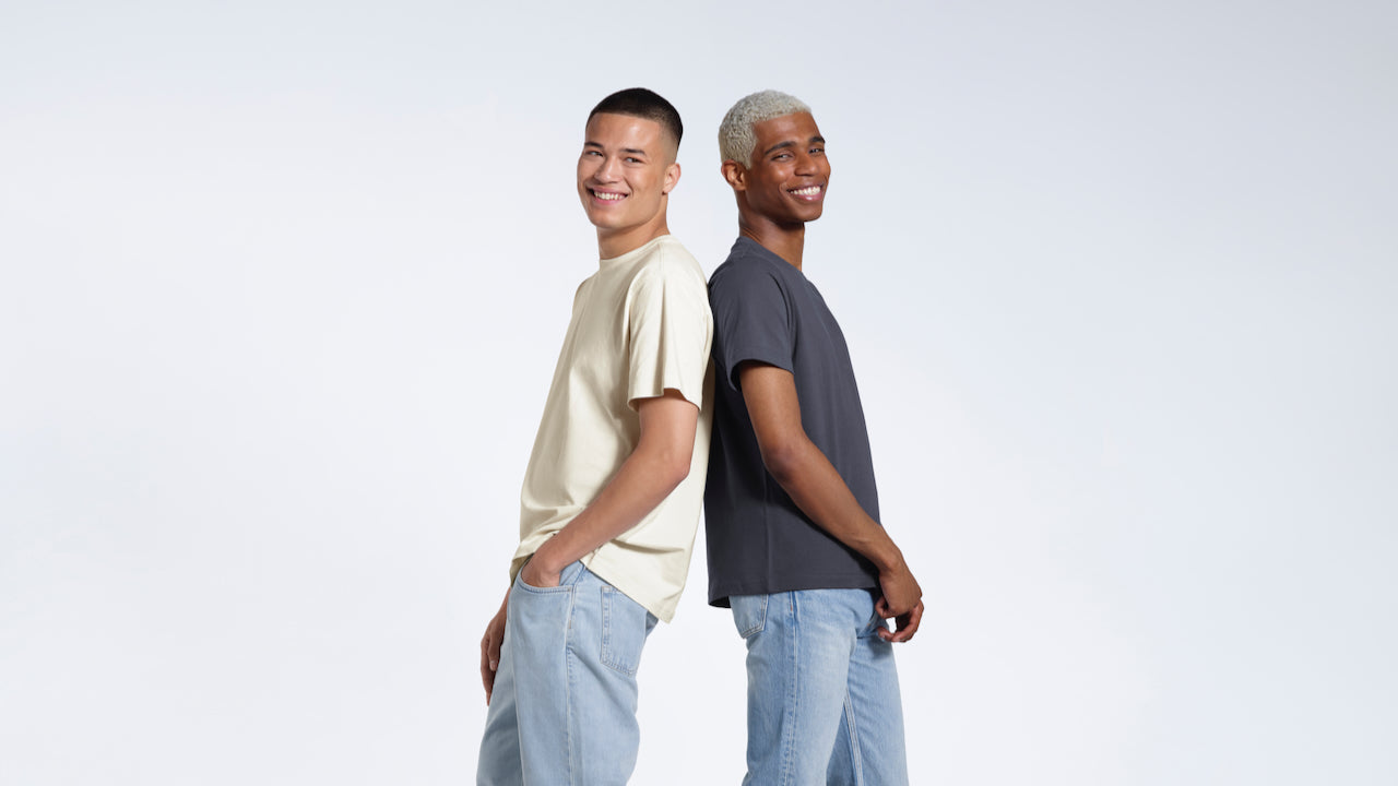 Two men standing back to back smiling, modelling plain ecru and dark grey coloured organic cotton t shirts.