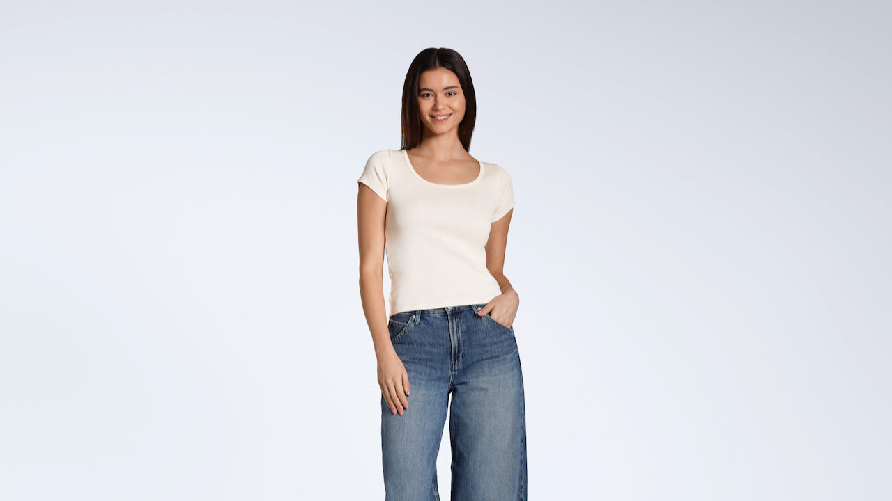 A woman smiling into the camera with her hand in her pocket, modelling a plain natural, organic cotton scoop neck t shirt.