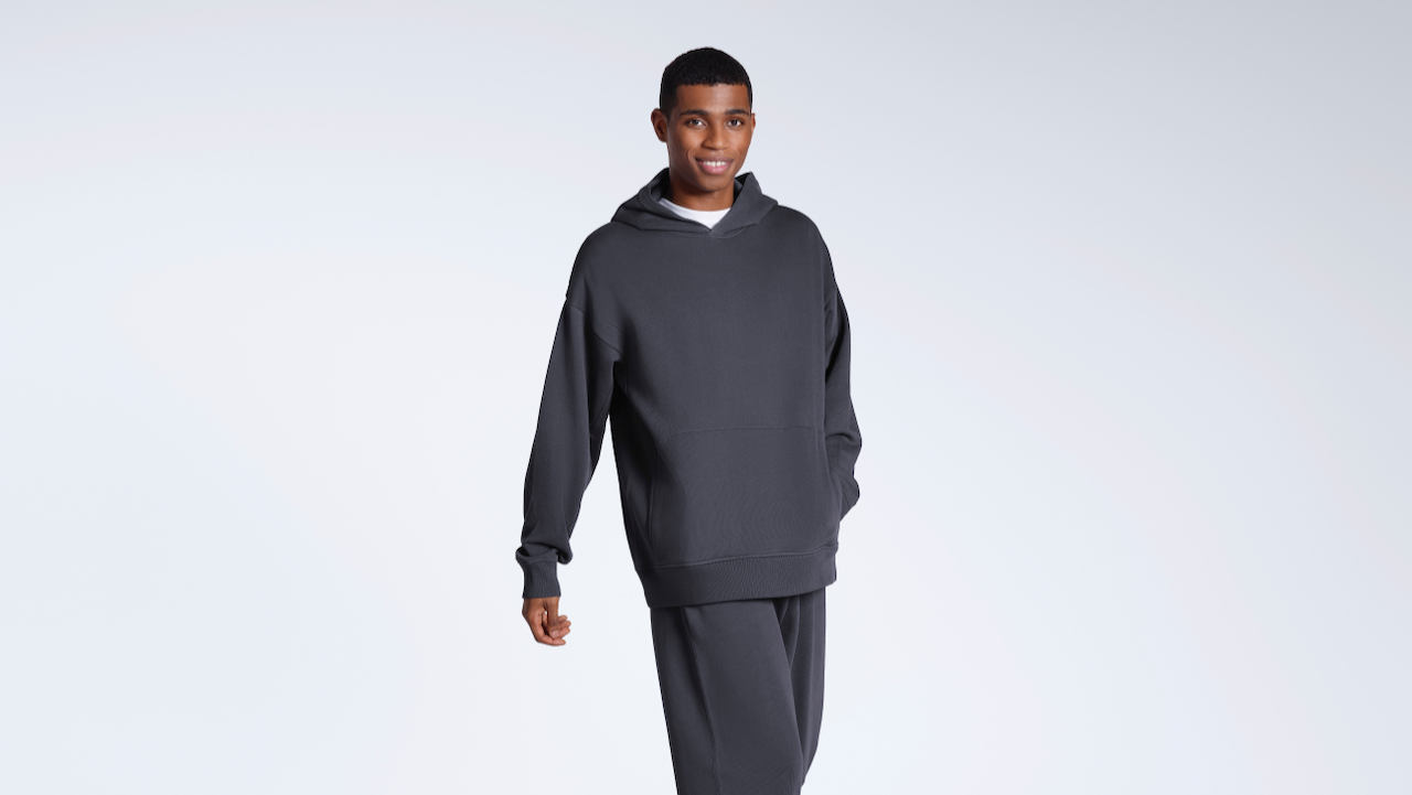 A man smiling into the camera with his hand in his pocket, modelling a plain dark grey, organic cotton hoodie with a matching pair of joggers.