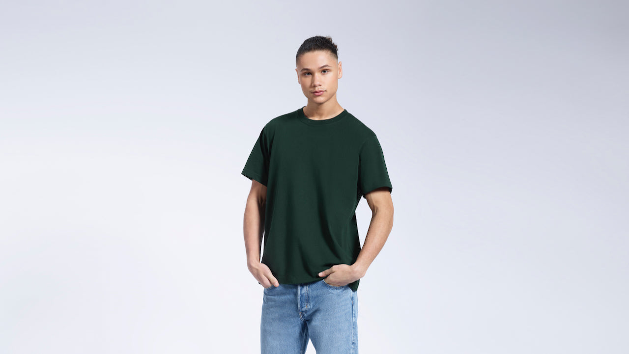 A man with his hands in his pockets, modelling a plain green short sleeve organic cotton t shirt.
