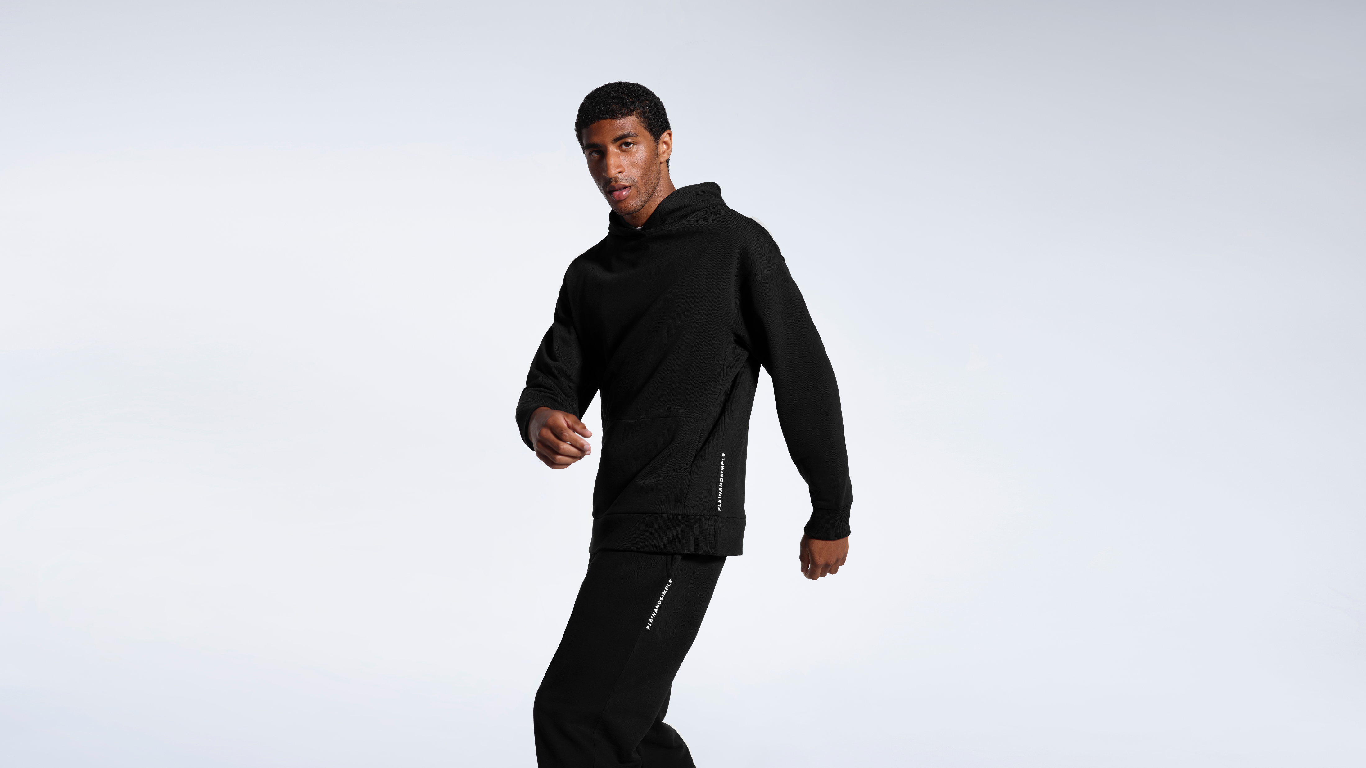 A looking at the camera turning, modelling a black heavyweight organic cotton traksuit.