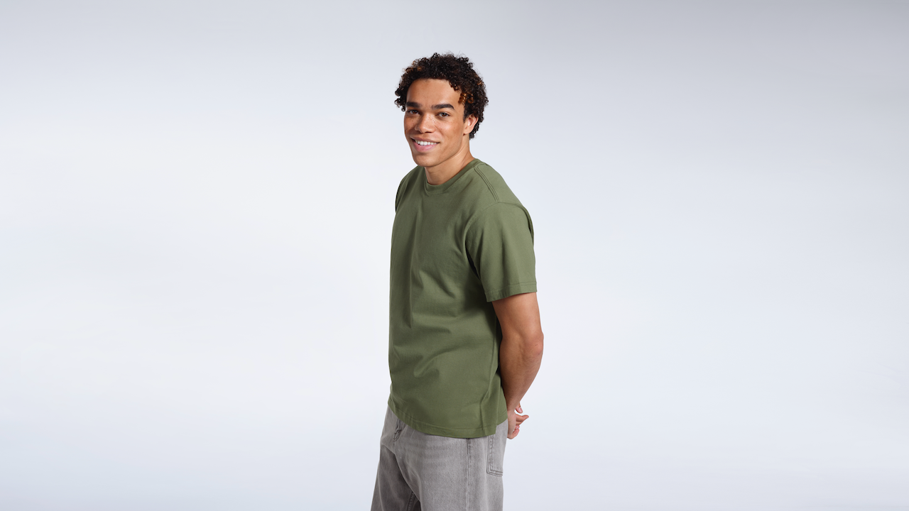 A man smiling into the camera with his hands behind his back, modelling a plain khaki green short sleeve organic cotton t shirt.