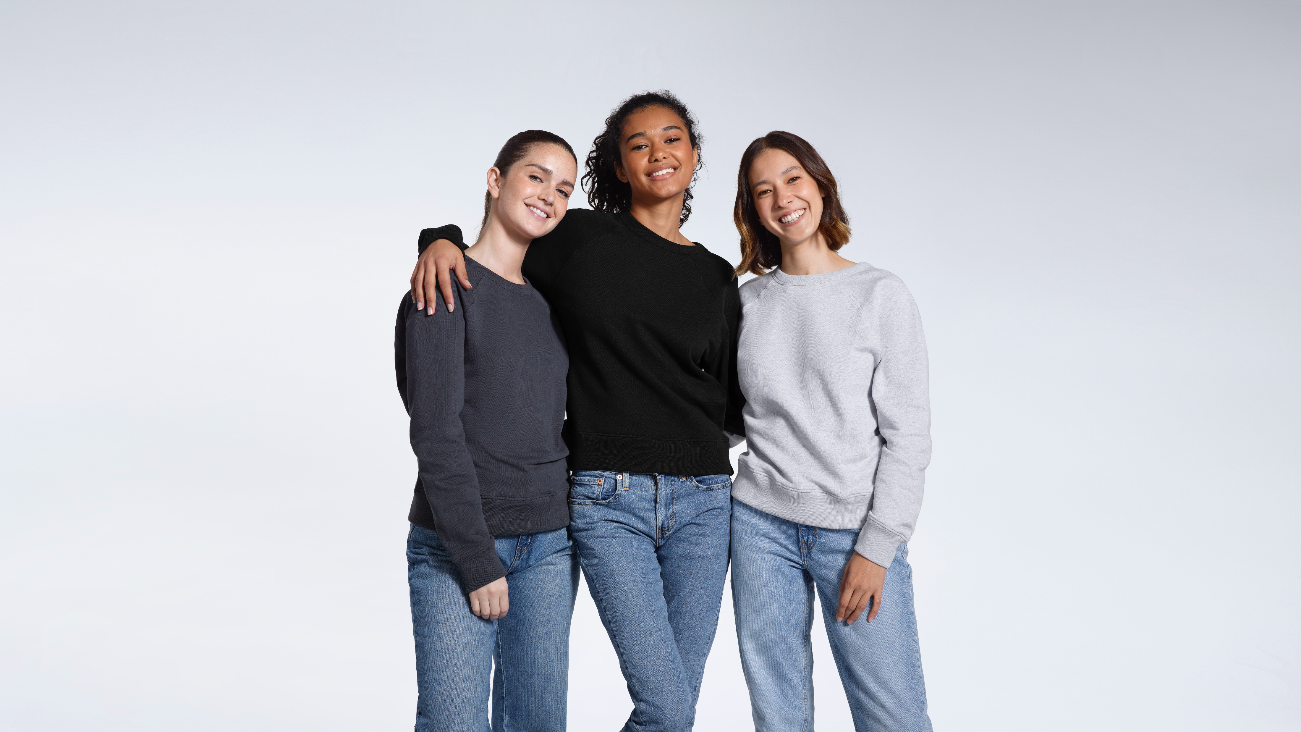 Three women smiling with their arms round each other, modelling plain organic cotton, dark grey, black and grey french terry sweatshirts.