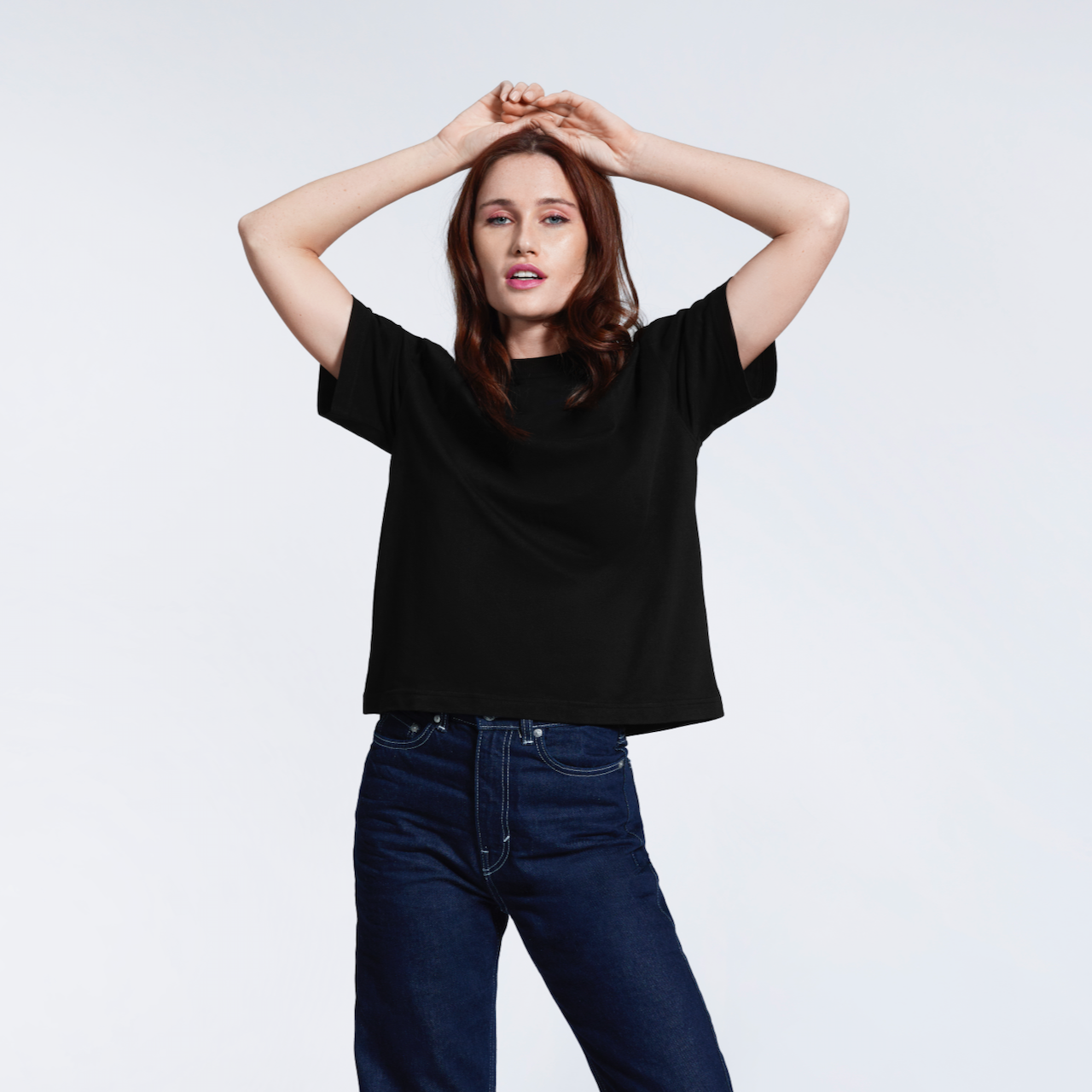 A woman with her hands on her head looking into the camera, modelling a plain black boxy, heavyweight organic cotton t shirt.