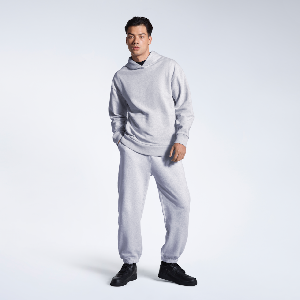 A man with his hand in his pocket, looking into the camera, modelling a matching grey, organic cotton hoodie with a pair of joggers.