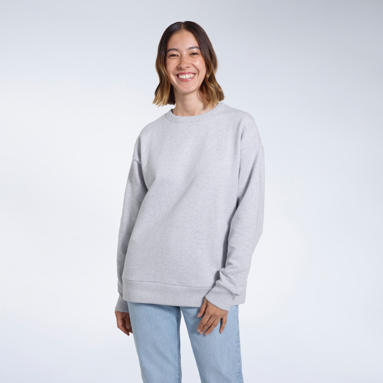 A woman smiling into the camera with her arms by her side, modelling a plain grey oversized organic cotton sweatshirt.