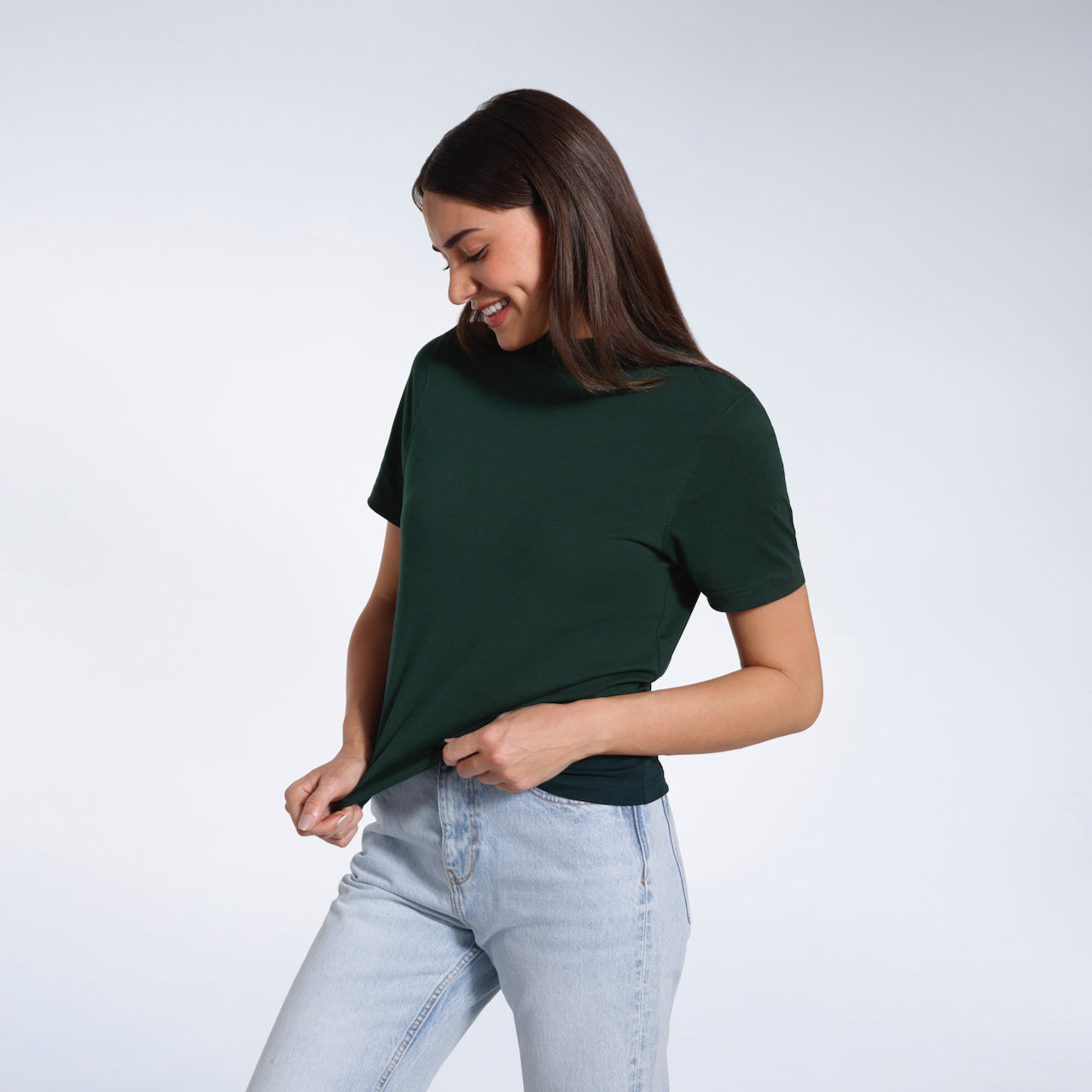 A woman looking down smiling and touching the bottom of her plain green short sleeve organic cotton t shirt.