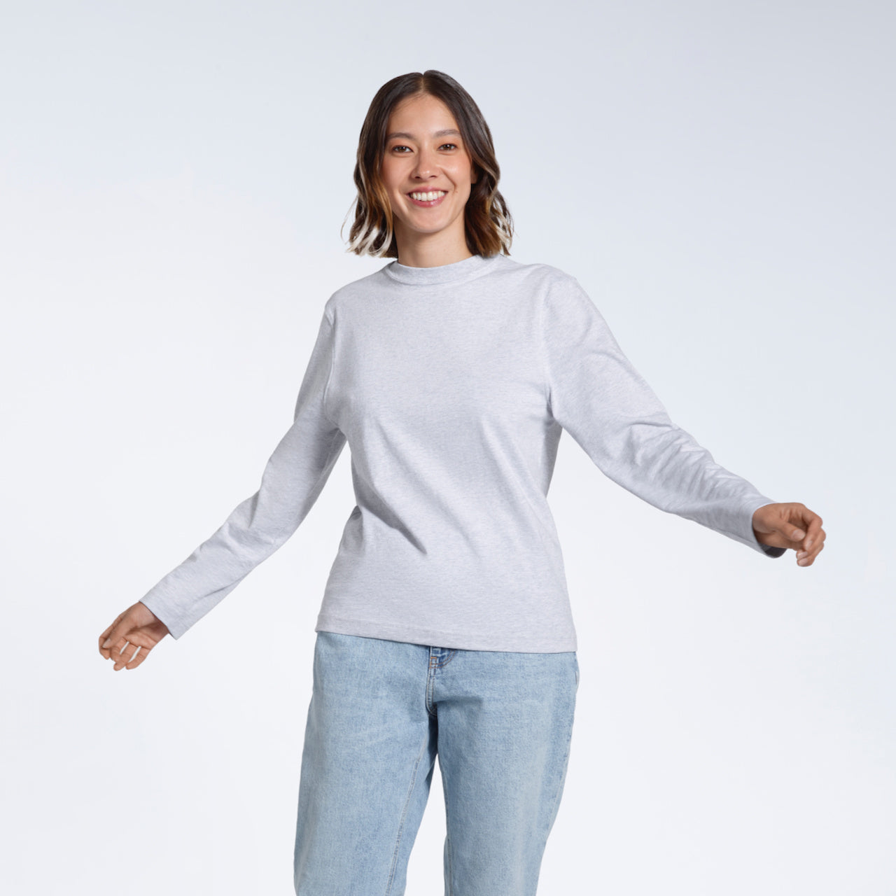 A woman smiling  into the camera with her arms out, modelling a plain grey long sleeve organic cotton t shirt.