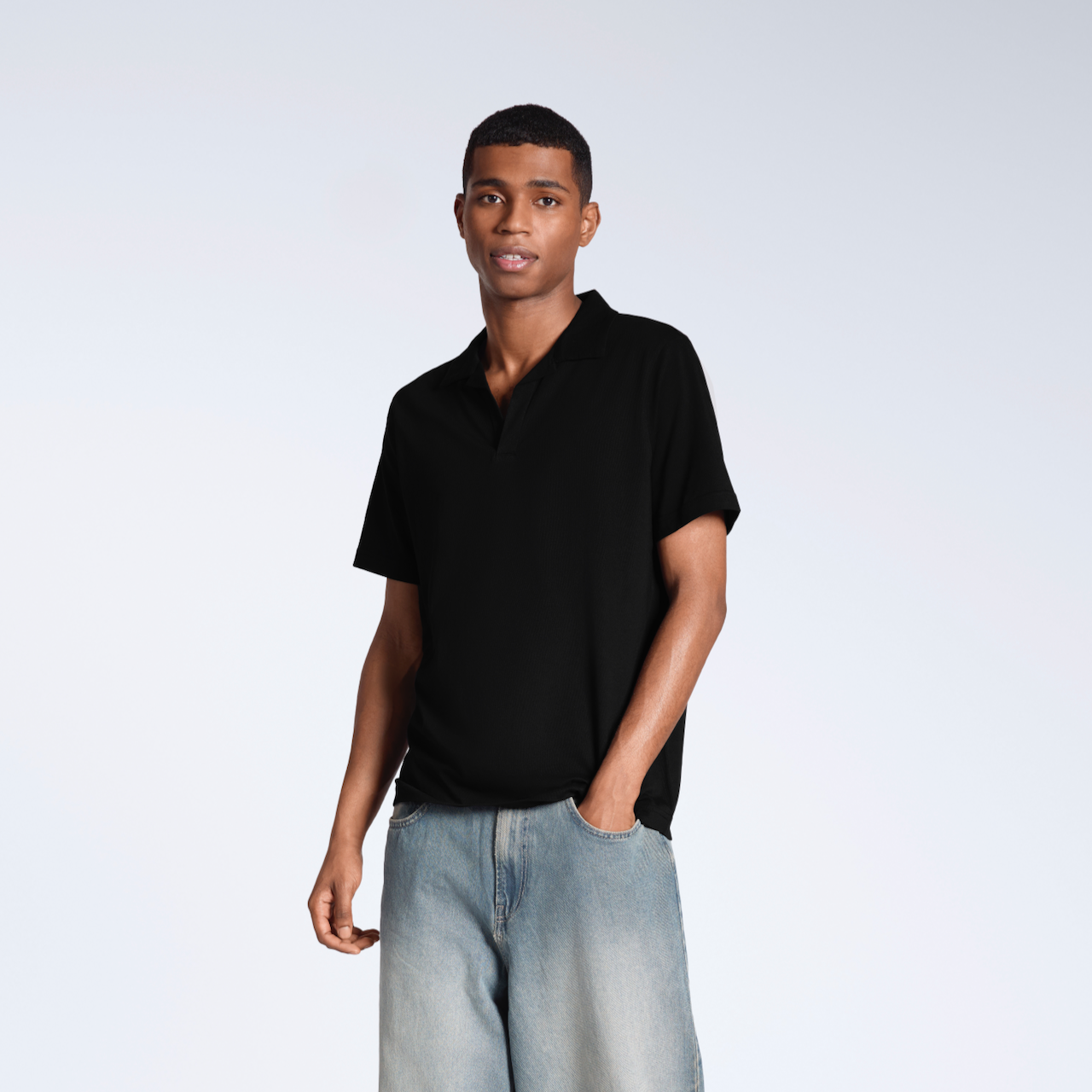 A man looking into the camera modelling a plain black, organic cotton, polo shirt with a pair of jeans.
