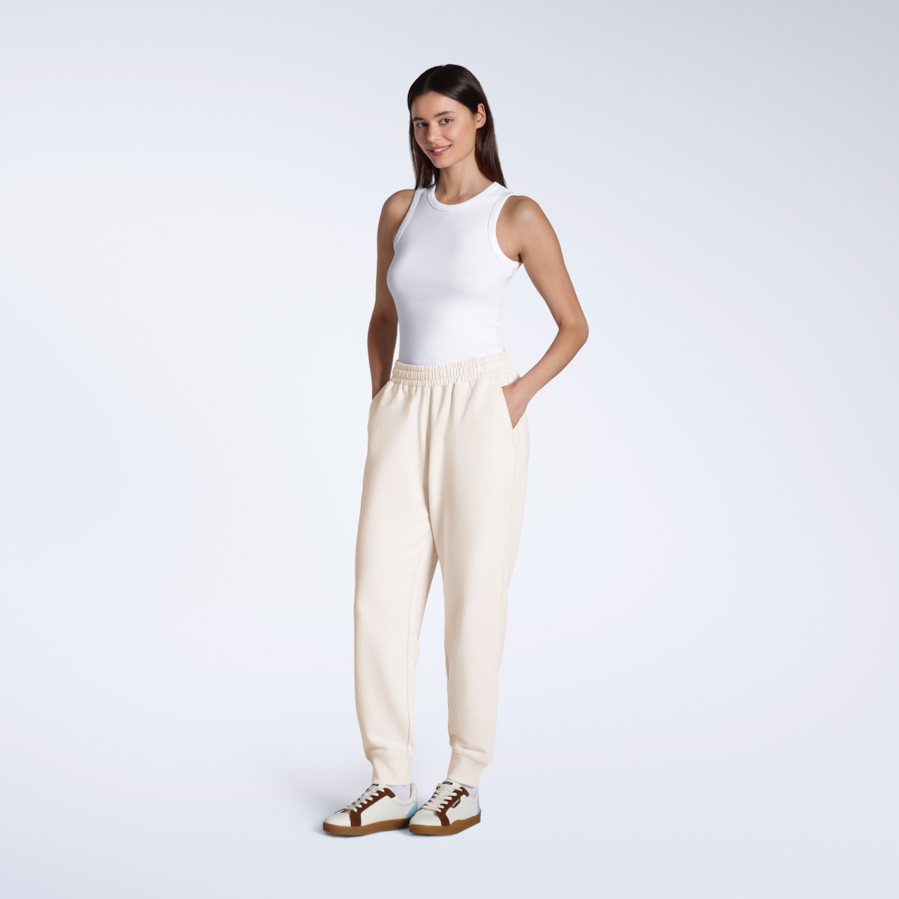 A full body photo woman with her hands in her pockets, modelling a pair of plain, natural coloured, organic cotton joggers.
