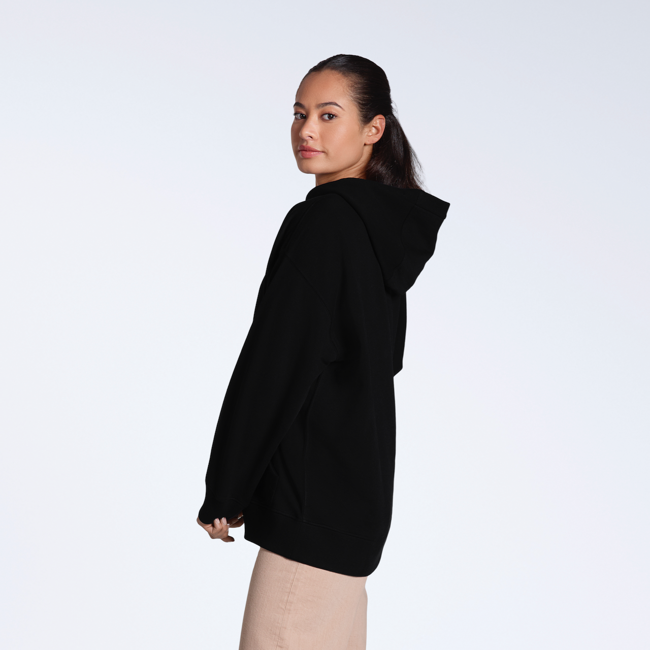 A woman facing side on looking back at the camera, modelling a plain black, oversized, organic cotton hoodie.