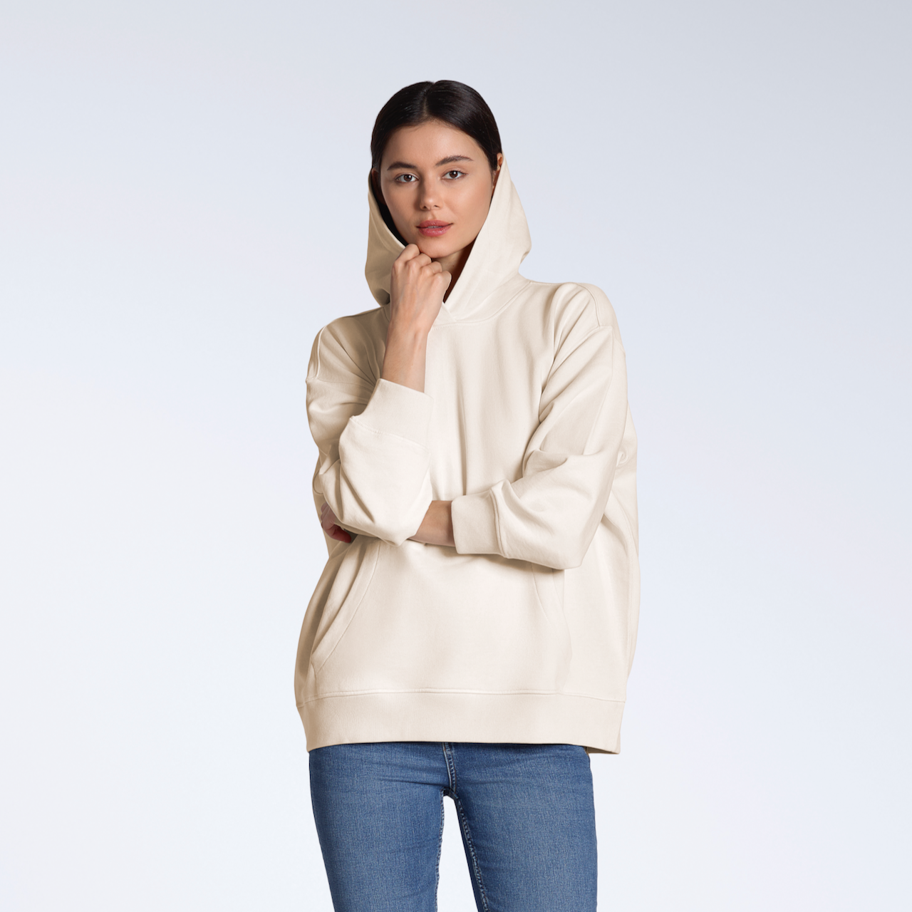 A woman looking into the camera touching her hood, modeling a plain, natural coloured, oversized, organic cotton hoodie.