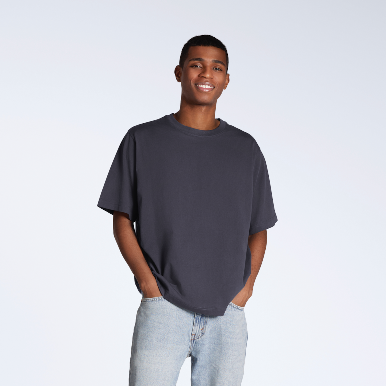 A man with his hands in his pockets smiling into the camera, modelling a plain dark grey, oversized, organic cotton t shirt.