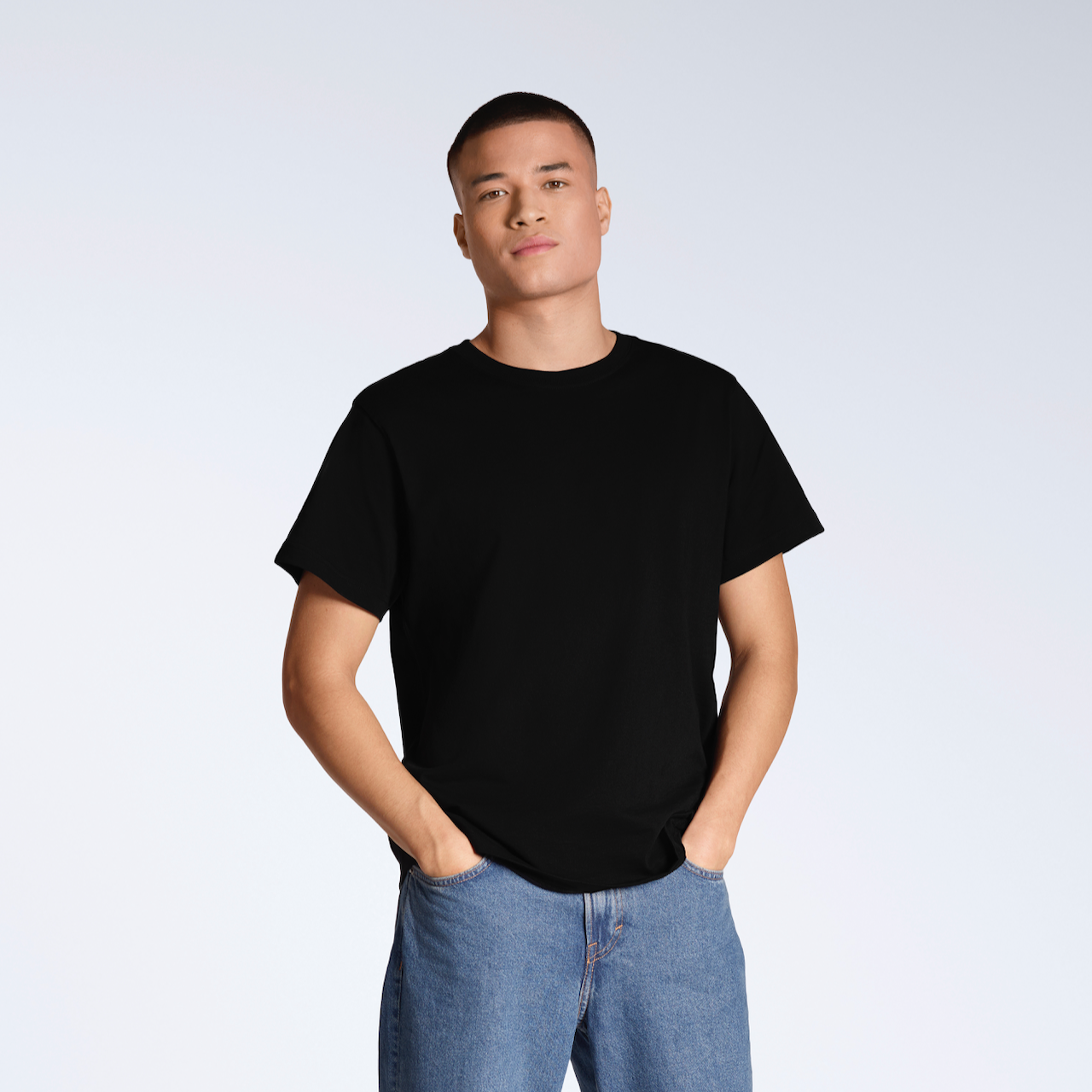 A man looking into the camera with his hands in his pockets, modelling a plain black, short sleeve organic cotton t shirt with a pair of jeans.
