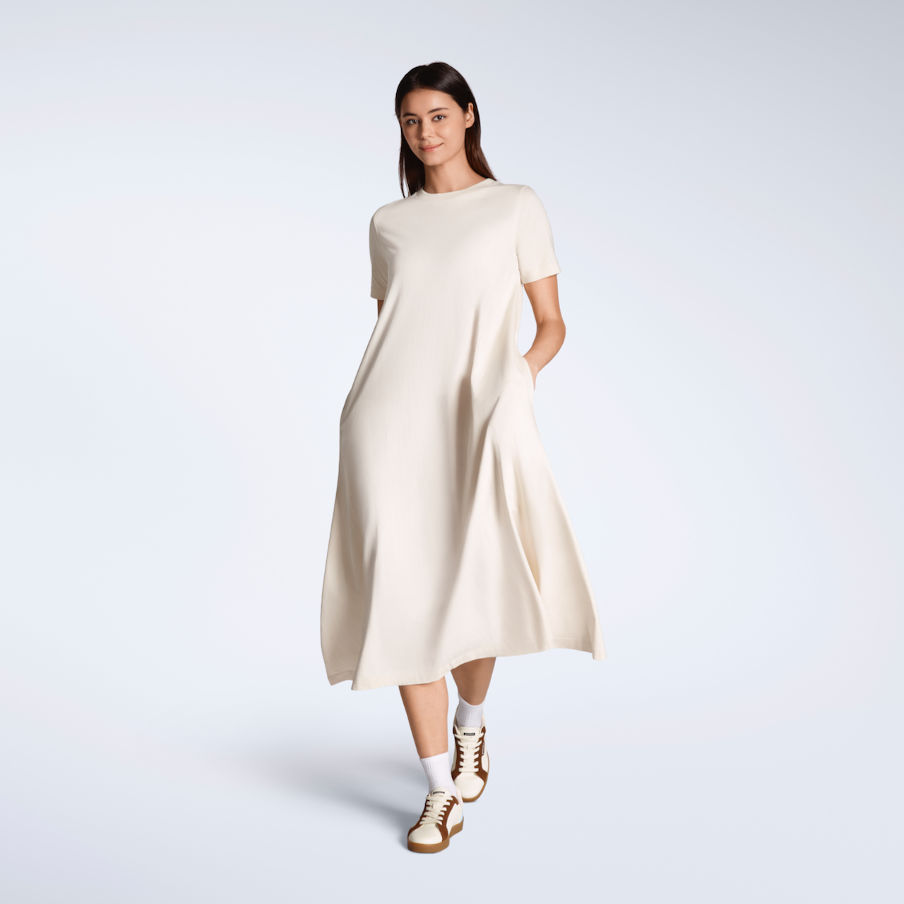 A woman smiling into the camera, walking, with her hands in her pockets modelling a plain natural coloured, long line, maxi organic cotton t shirt dress.