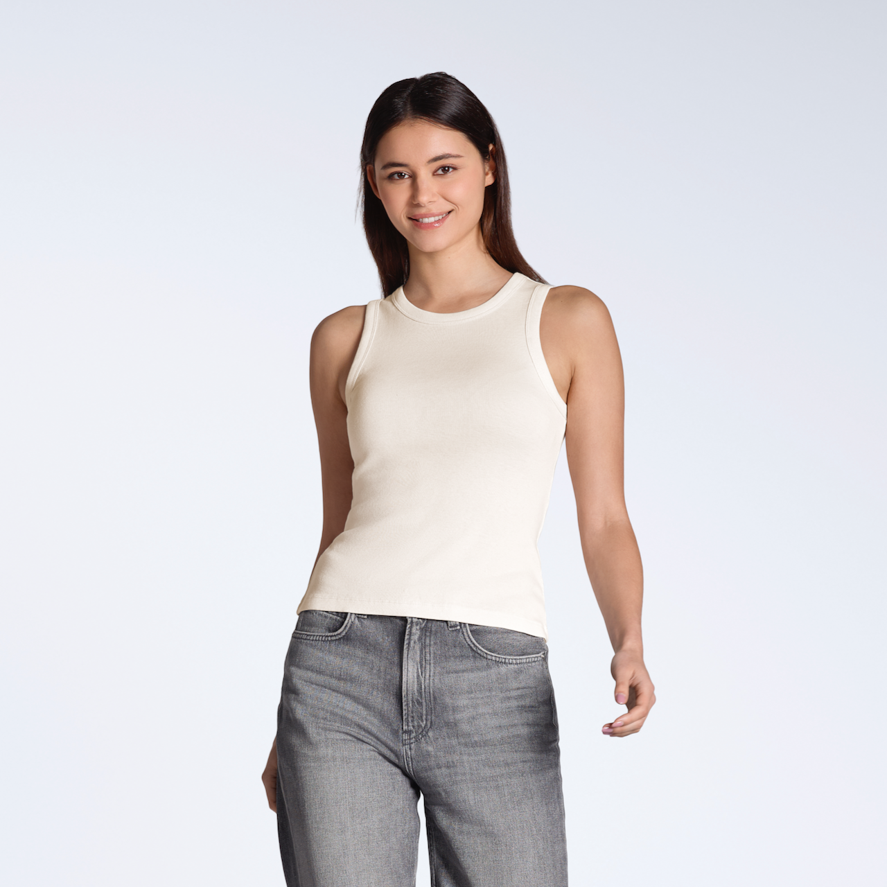 A woman smiling into the camera, modelling a plain natural coloured, organic cotton tank top with a pair of jeans.