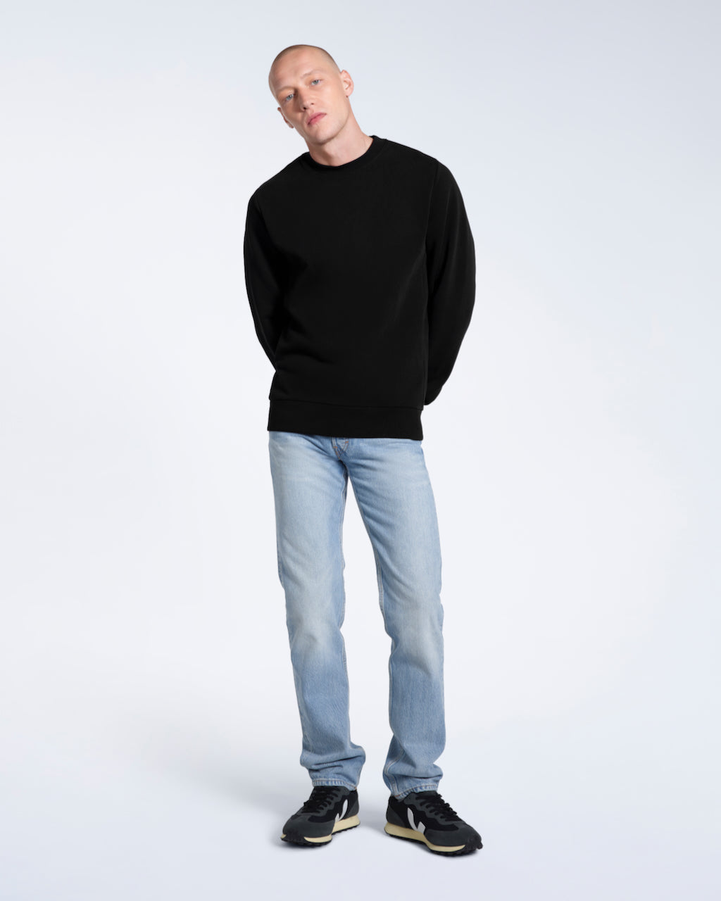 A full body photo of a man looking into the camera with his arms behind his back, modelling a plain black french terry organic cotton sweatshirt.