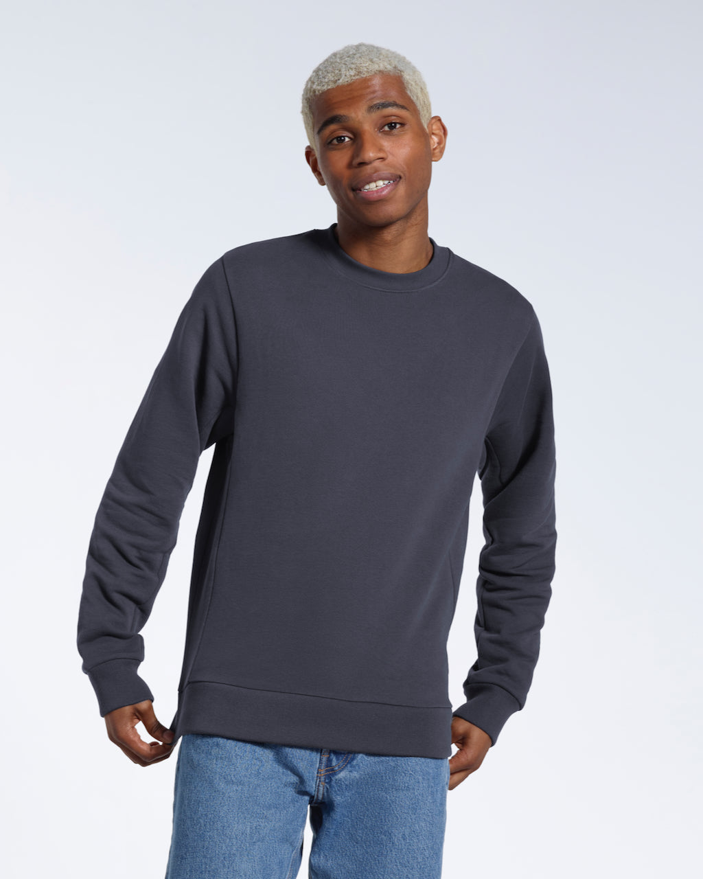 A man smiling into the camera, touching the bottom of his plain dark grey french terry organic cotton sweatshirt.