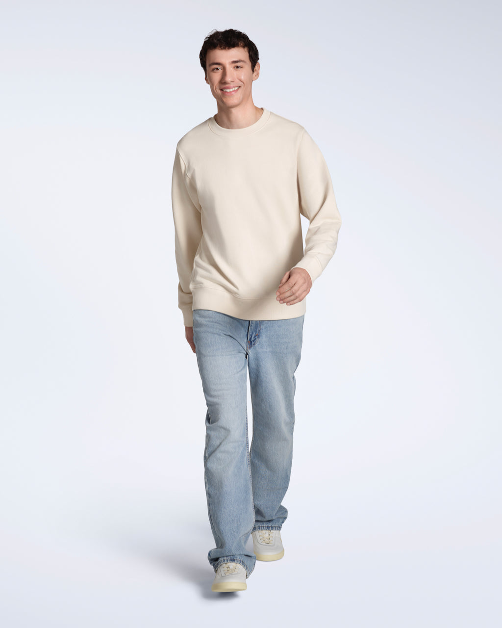 A full body photo of a man smiling walking towards the camera, modelling a plain natural coloured, organic cotton sweatshirt with a pair of blue jeans.
