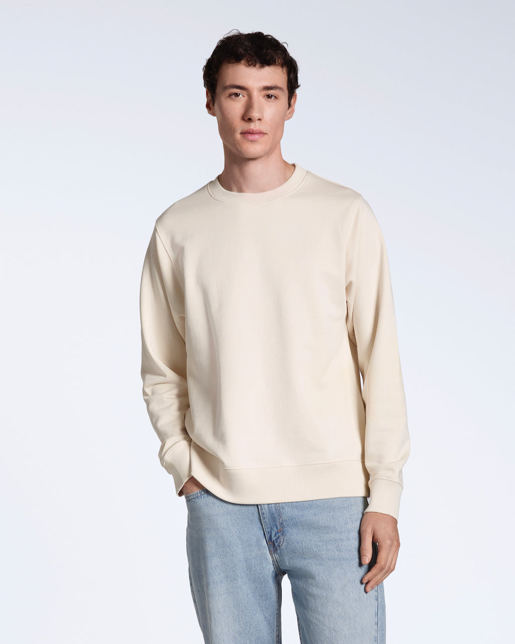 A man with his hand in his pocket looking into the camera, modelling a plain natural coloured, organic cotton sweatshirt with a pair of blue jeans.