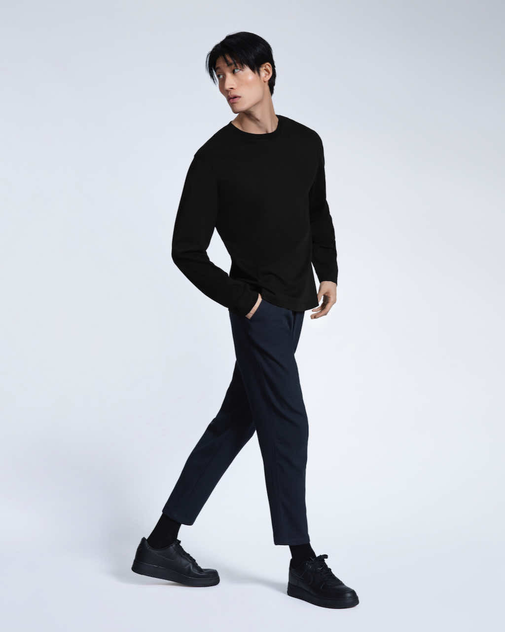 A full body photo of a man walking with his hand in his pocket, modelling a plain black long sleeve organic cotton t shirt.