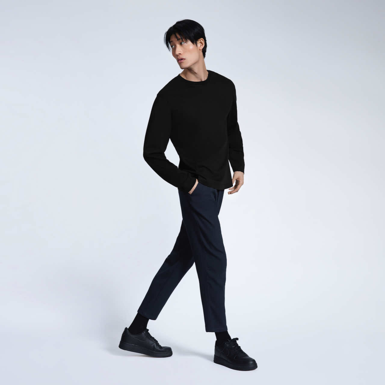 A full body photo of a man walking with his hand in his pocket, modelling a plain black long sleeve organic cotton t shirt.