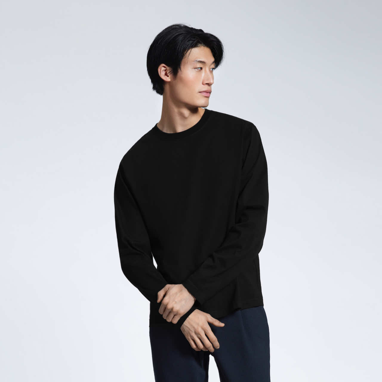 A man with his hands crossed looking away from the camera, modelling a plain black long sleeve organic cotton t shirt.