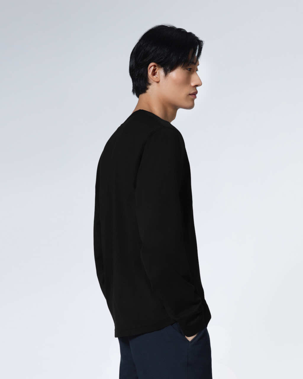 A man facing side on with his hands in his pockets, modelling a plain black long sleeve organic cotton t shirt.