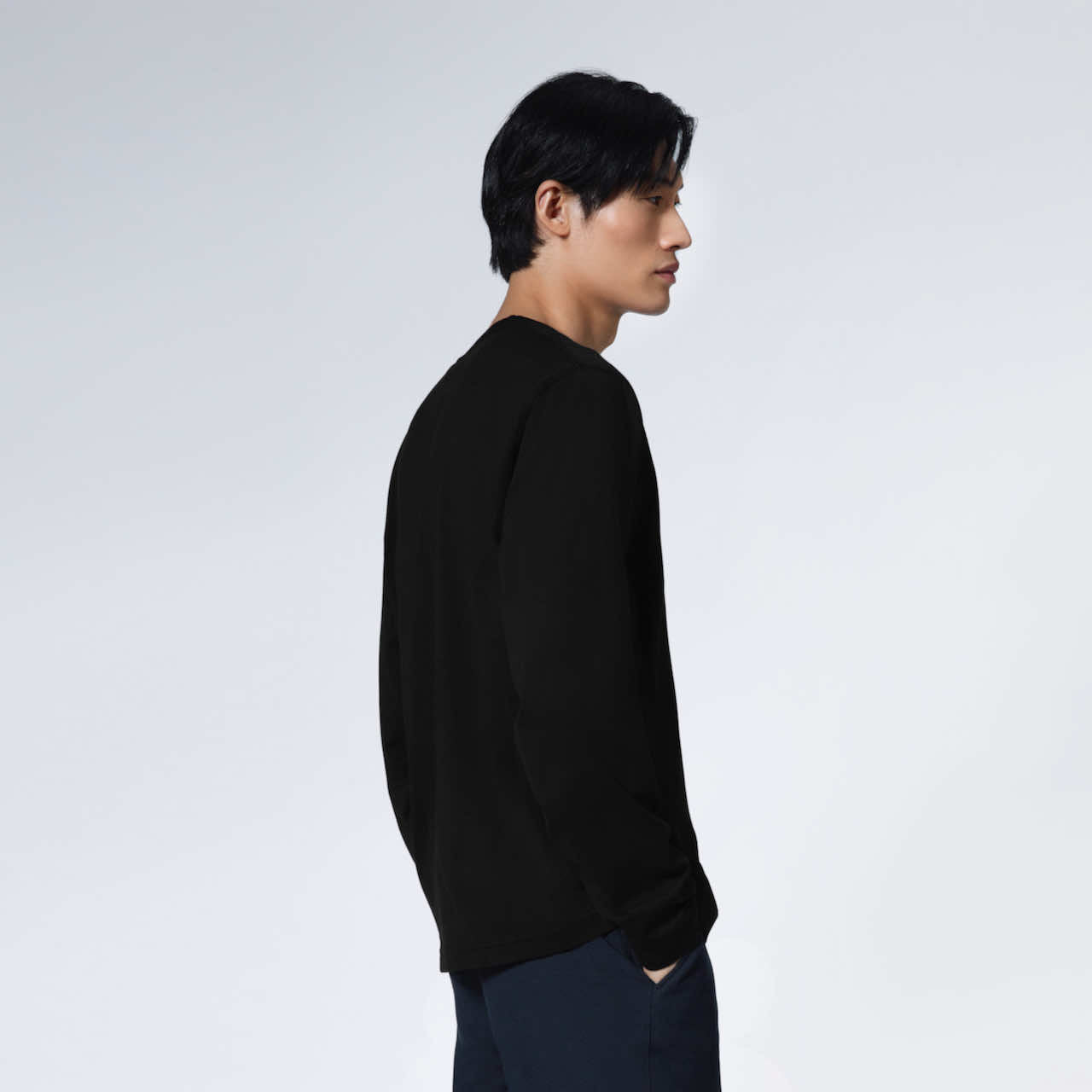 A man facing side on with his hands in his pockets, modelling a plain black long sleeve organic cotton t shirt.
