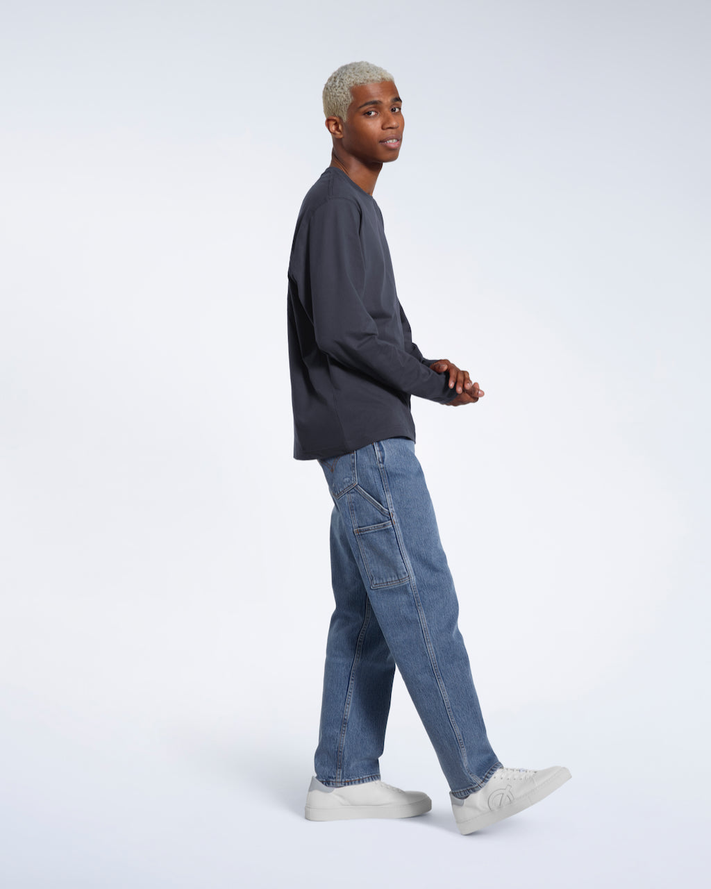 A full body photo of a man side on looking into the camera, modelling a plain dark grey long sleeve organic cotton t shirt.