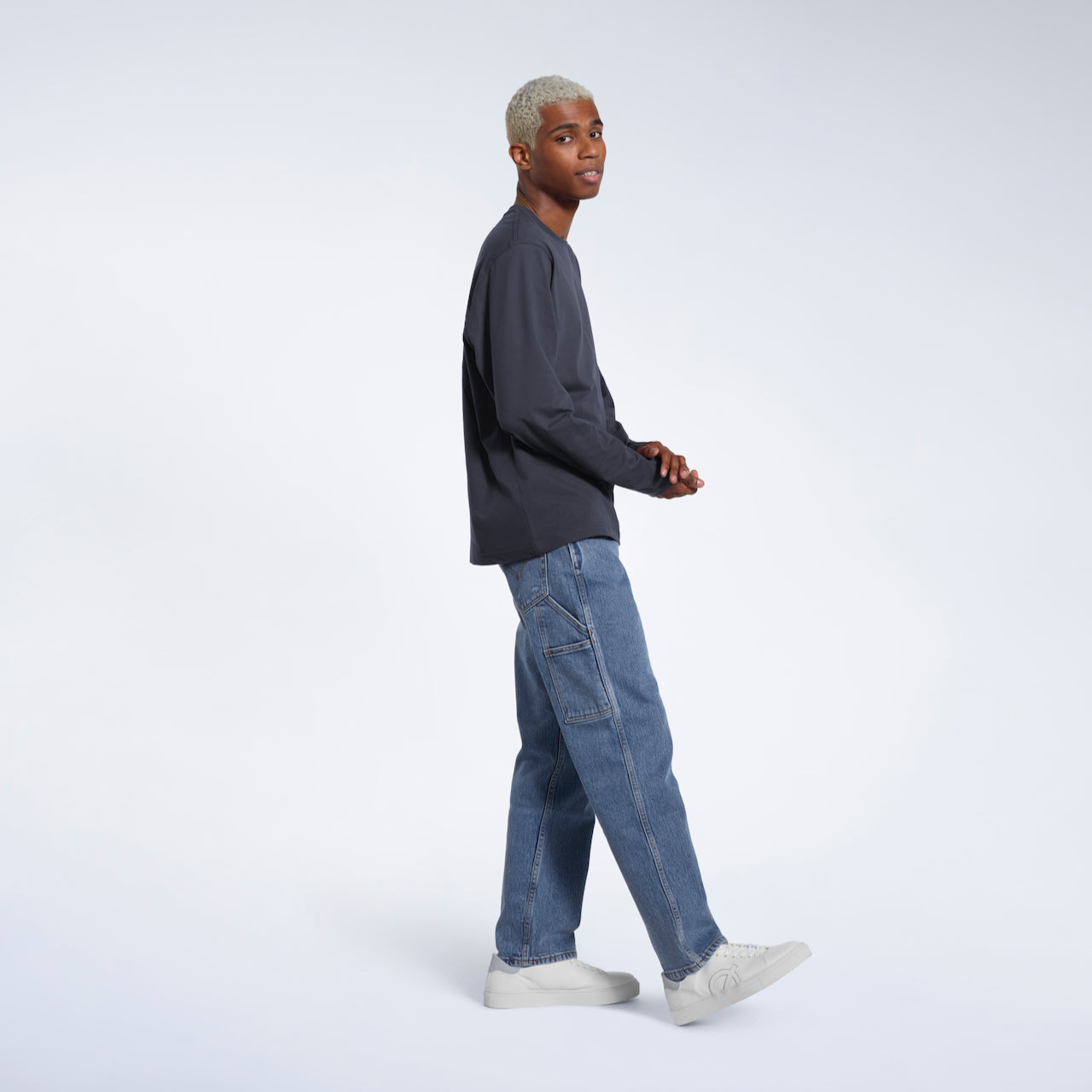 A full body photo of a man side on looking into the camera, modelling a plain dark grey long sleeve organic cotton t shirt.
