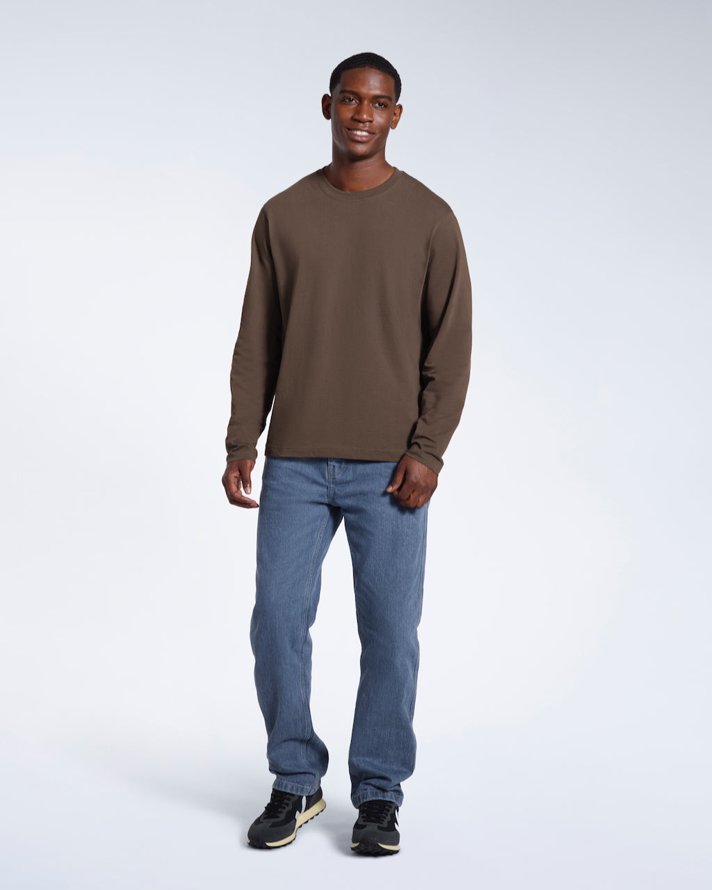 A full body photo of a man smiling, modelling a plain dark khaki long sleeve organic cotton t shirt.