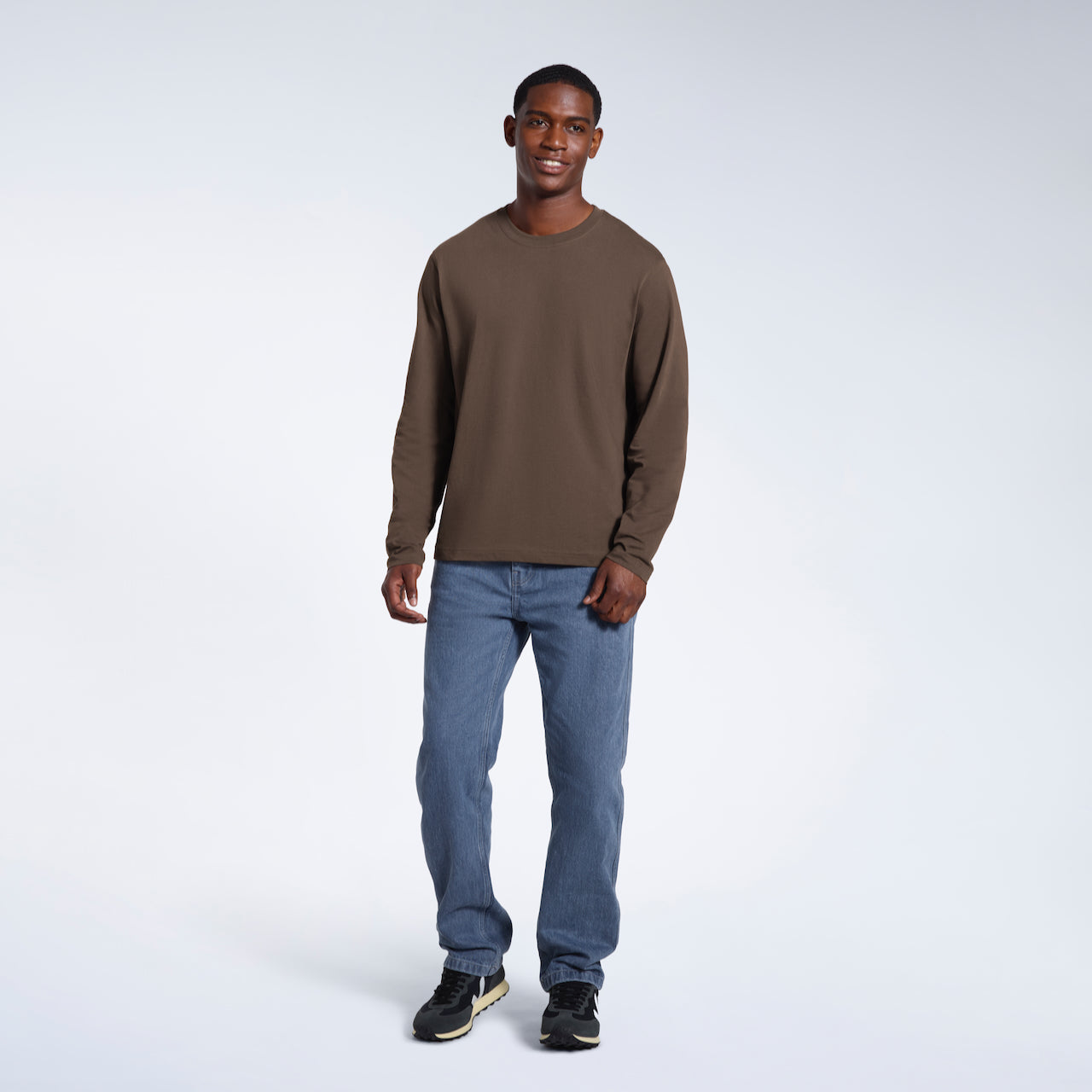 A full body photo of a man smiling, modelling a plain dark khaki long sleeve organic cotton t shirt.