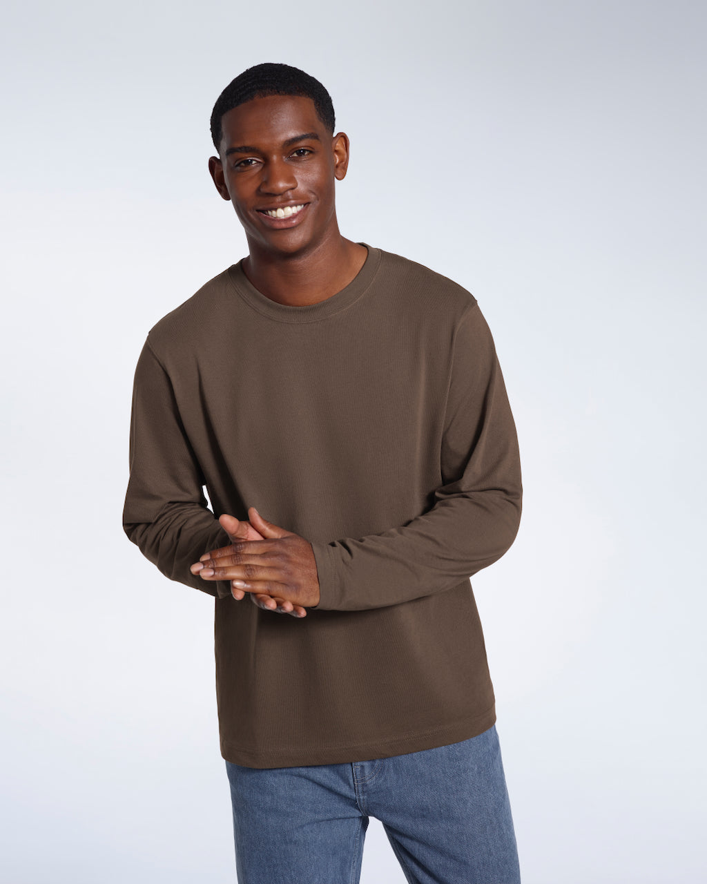 A man smiling into the camera with his hands together, modelling a plain dark khaki long sleeve organic cotton t shirt.